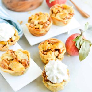 Square, close up image of mini apple pie tarts topped with whipped cream on a variety of white serving dishes.