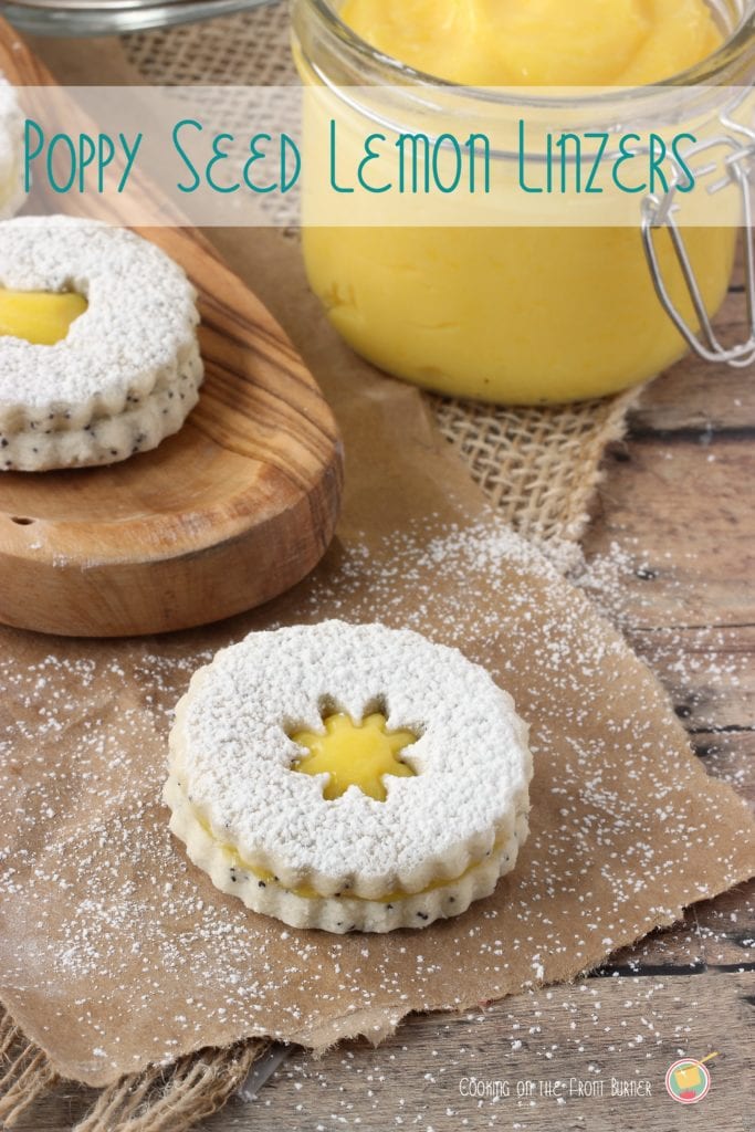 Two lemon poppy seed linzer cookies sit on top of a brown background. Each is sprinkled with powdered sugar. In the background, there is a clear jar filled with lemon curd.