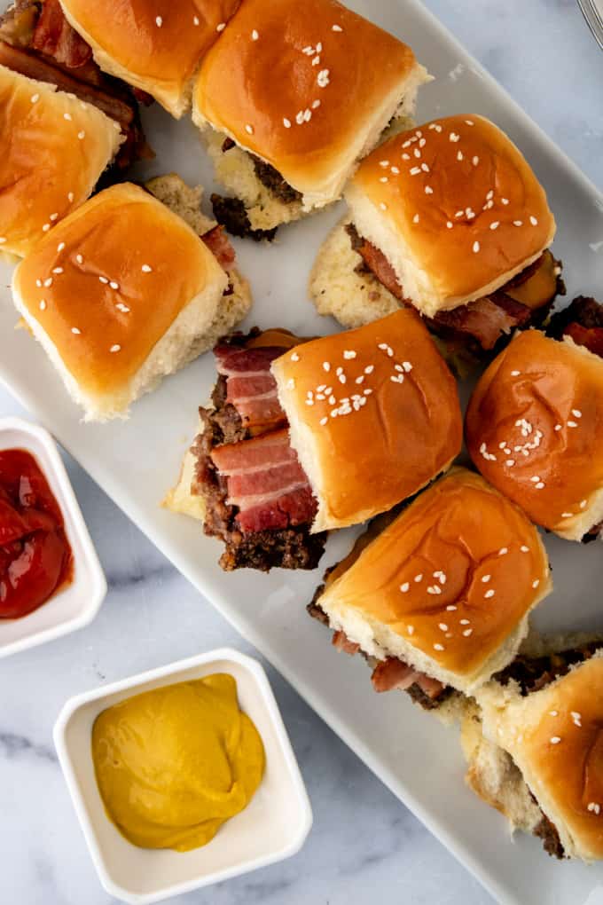 Top down view of bacon cheeseburger sliders on a white plate next to small bowls filled with ketchup and mustard.