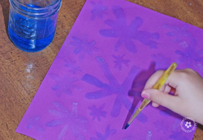 Child painting blue snowflakes on a purple piece of paper.