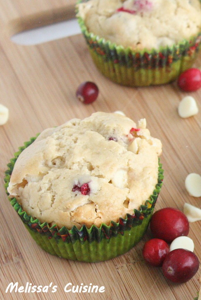 Close up image of Cranberry and White Chocolate Muffins on a wood counter top, with fresh cranberries and white chocolate chips.