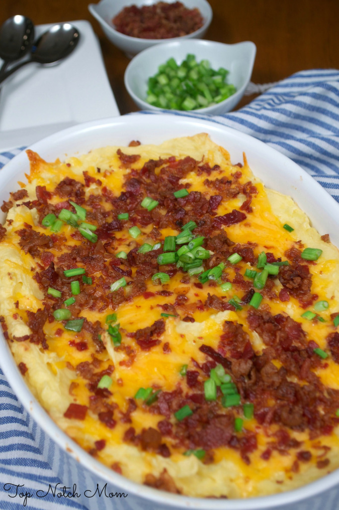 Baked Potato Casserole with yellow cheddar cheese, bacon and green onions on a white plate with bowls of green onions and bacon in the background.