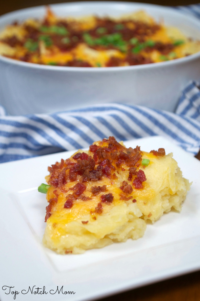 Baked Potato Casserole with yellow cheddar cheese, bacon and green onions on a white plate with the casserole dish in the background.