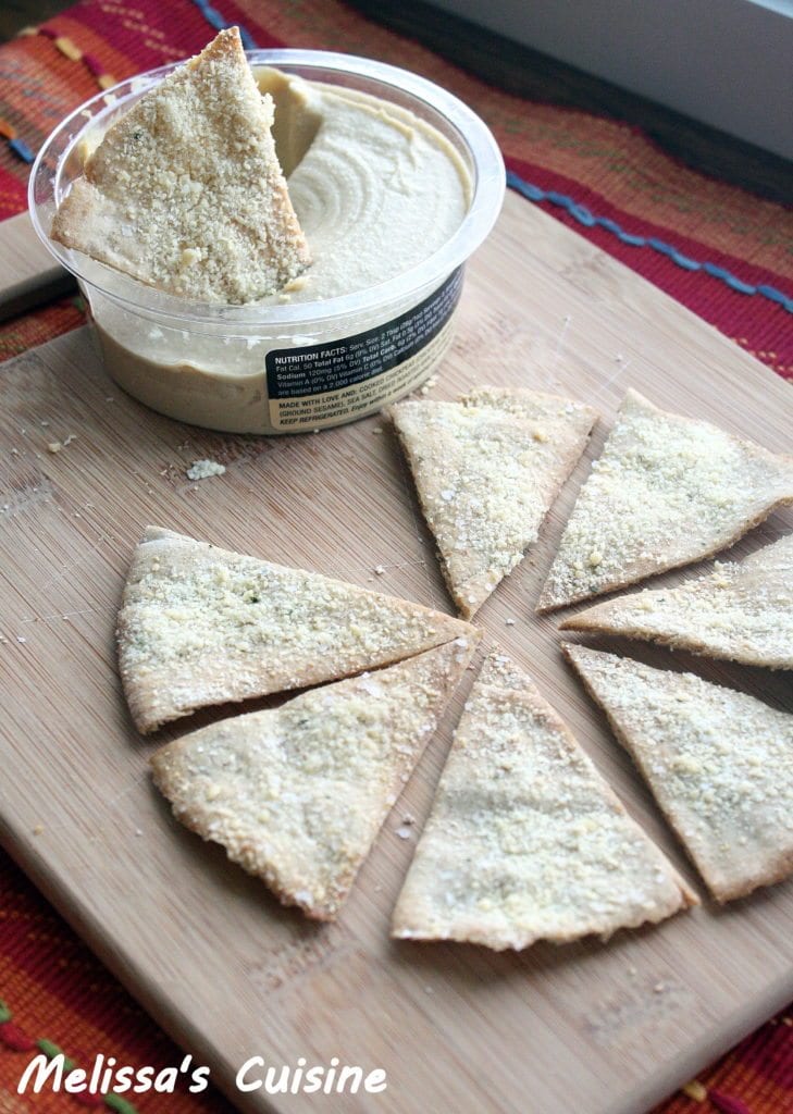 Pita chips laid out on a wood cutting board with a container of hummus behind it with a pita chip sticking out of it.