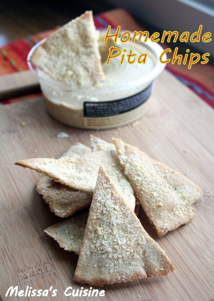 Pita chips sprinkled with parmesan cheese stacked in a pile on a wood cutting board with a container of hummus in the back with a pita chip dipped into it.