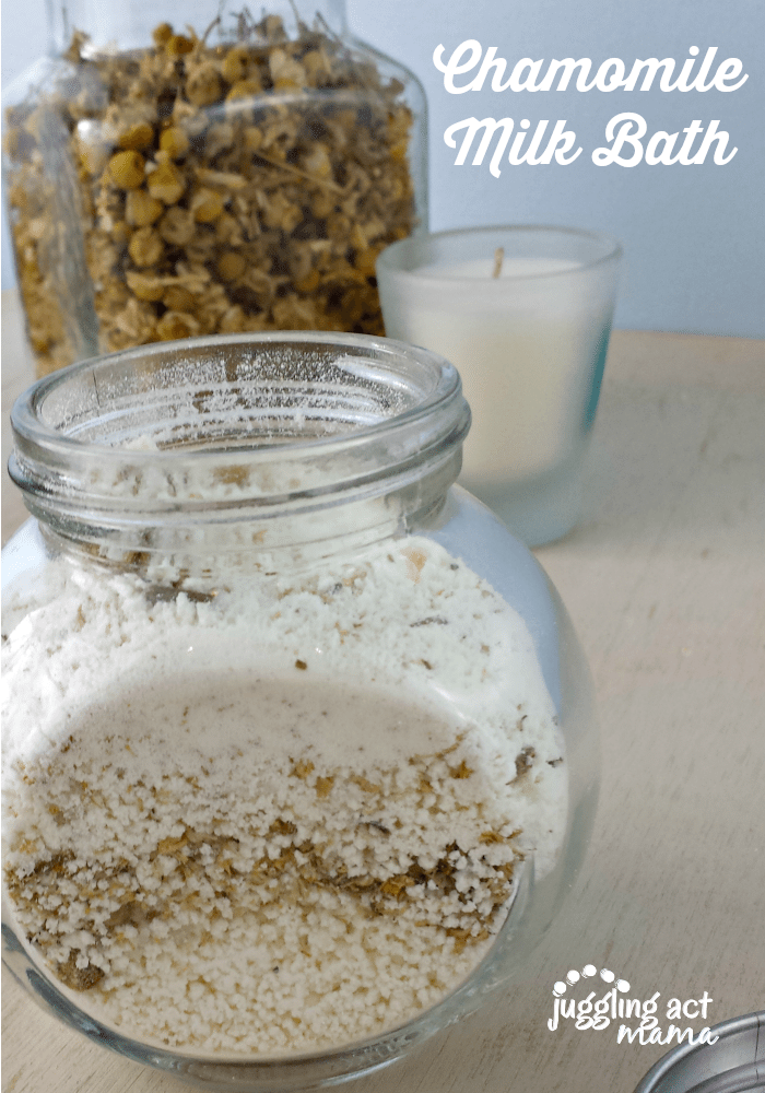 Close up image of a small glass jar filled with chamomile milk bath in front of a candle and jar of dried chamomile.