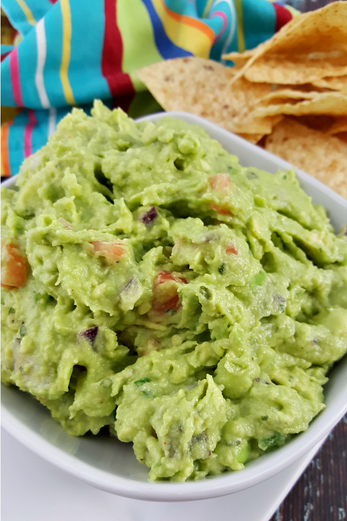 A close up of a bowl of homemade guacamole with tomatoes and red onions next to tortilla chips and a festive kitchen towel.