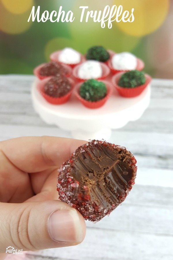 Hand holding a mocha truffle in front of pretty mocha truffles on a white cake stand.