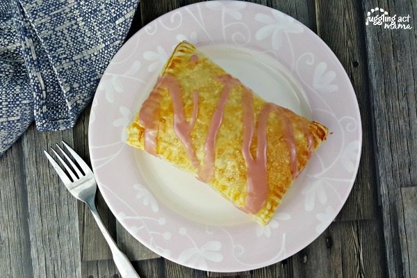 Arial view of a strawberry cream cheese danish on a white plate with pink icing drizzled on top with a fork beside the plate and blue napkin.