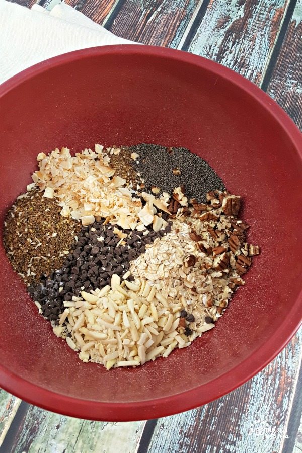 A red mixing bowl sits on a wooden table. The bowl is full of dry ingredients for making No Bake Chocolate Chip Peanut Butter Energy Bites, including toasted coconut, rolled oats, slivered almonds, chia seeds, mini chocolate chips, and chopped pecans.