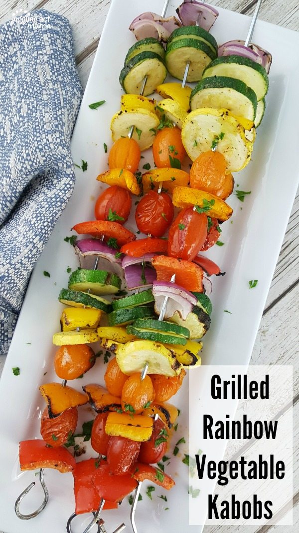 Grilled Vegetable Kabobs with tomatoes, zucchini, summer squash, red onions, plus red, orange, and yellow peppers on metal skewers piled high on a white serving tray next to a blue towel.
