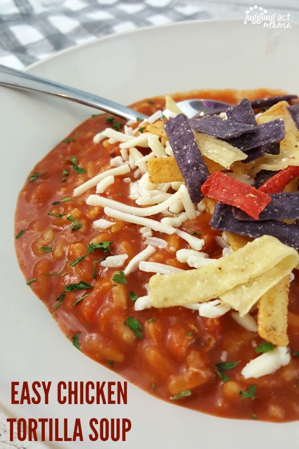A close up of a white wide-mouth bowl of easy chicken tortilla soup with tri-color tortilla and shreaded cheese sits on wooden planks, with a white and gray napkin nearby. A spoon is in the bowl and the soup is garnished with parsley as well.