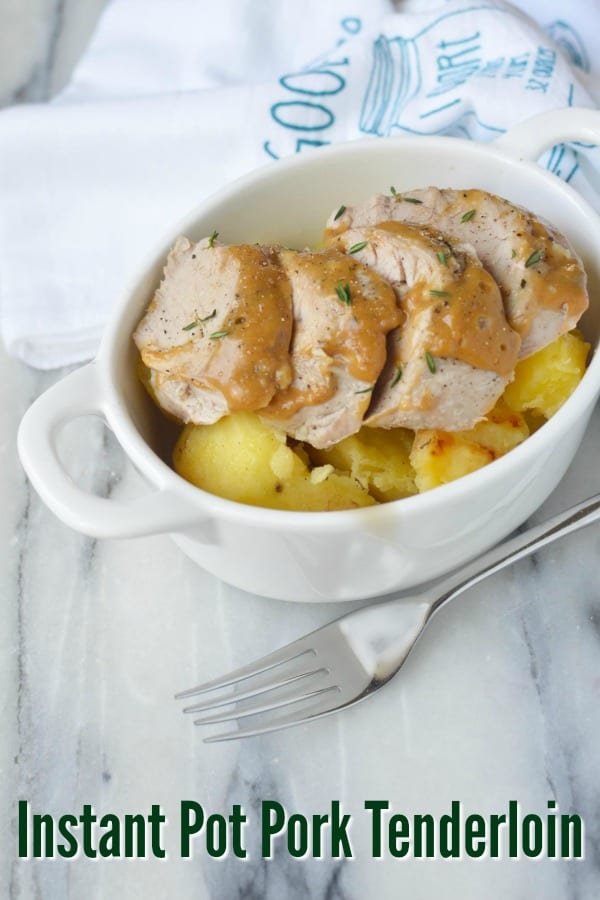 Sliced Instant Pot Pork Tenderloin on top of potatoes in an oval white dish with handles with a silver fork and a blue and white towel in the background.