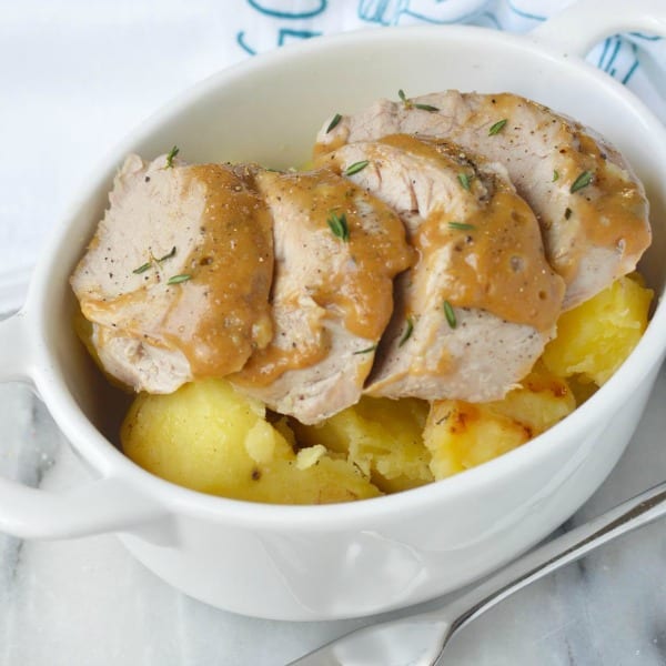 Close up of sliced Instant Pot Pork Tenderloin on top of potatoes in an oval white dish with handles with a silver fork and a blue and white towel in the background.