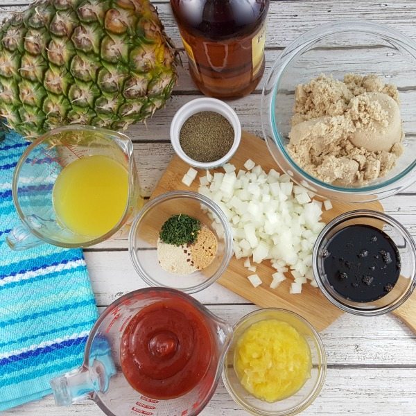 Ingredients for Pineapple Bourbon BBQ Sauce are picutred on a white table with a blue cloth, including a measuring cup with pineapple juice and another one with ketchup, bottle of bourbon, whole pineapple, a small cutting board with diced onion and small bowls of light brown sugar, black pepper, and crushed pineapple.