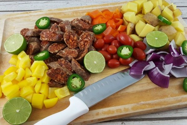 Raw vegetables and chiorizo on a wooden cutting board with a knife.