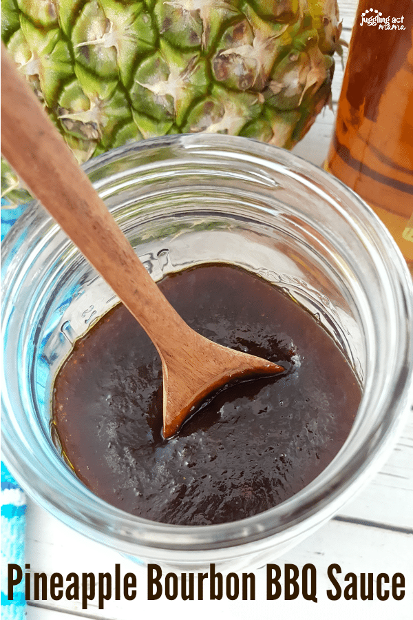 A mason jar of Pineapple Bourbon BBQ Sauce with a wooden spoon in it. In the background are a whole pineapple and a bottle of bouron.