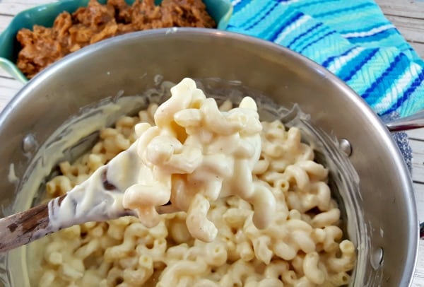 A spoonful of mac and cheese scooped from a full stock potwith a dish of pulled pork in the background next to a turqoise towel.