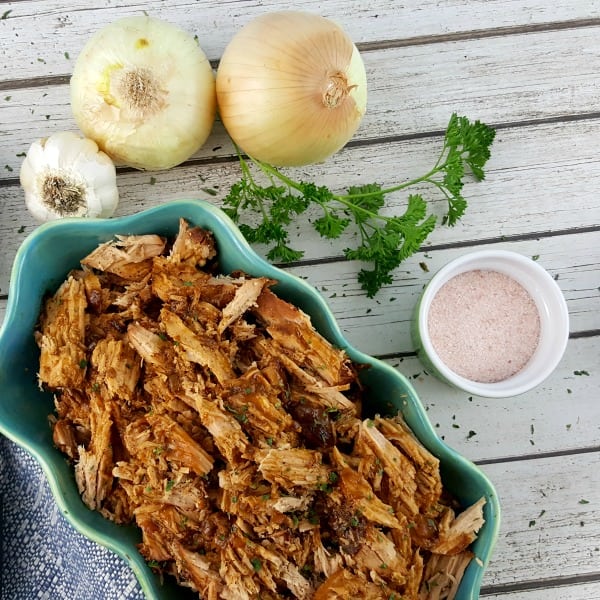 Pulled pork in an oval serving dish surrounded by garlic, onions, fresh herbs and salt.