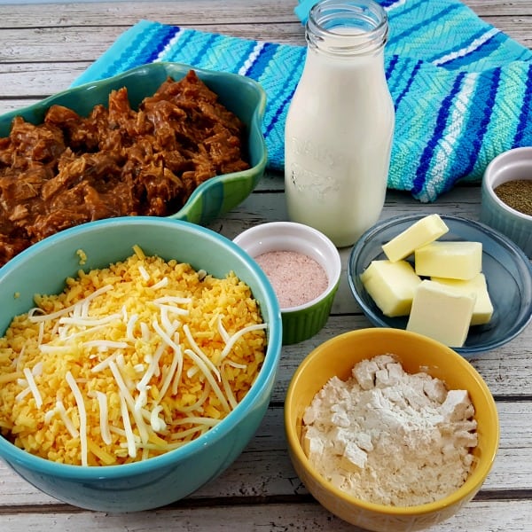 Ingredients to make Pulled Pork Mac and Cheese sit on a table with a turquoise kitchen towel. Ingredients include cheese in a light blue bowl, an oval dish of pulled pork, butter, cream, salt and pepper.