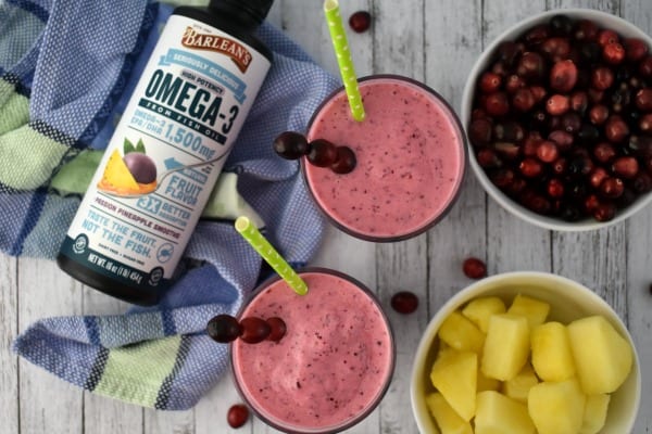 Two pineapple cranberry smoothies with green straws on a wooden table next to a blue hand towel with a bottle of Barlean's omega swirl
