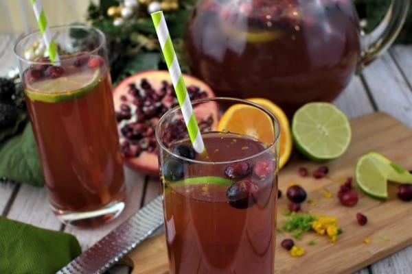 close up of Pomegranate Punch in a glass pitcher along with tall glasses of punch garnished with fresh limes, oranges, cranberries and pomegranate seeds.