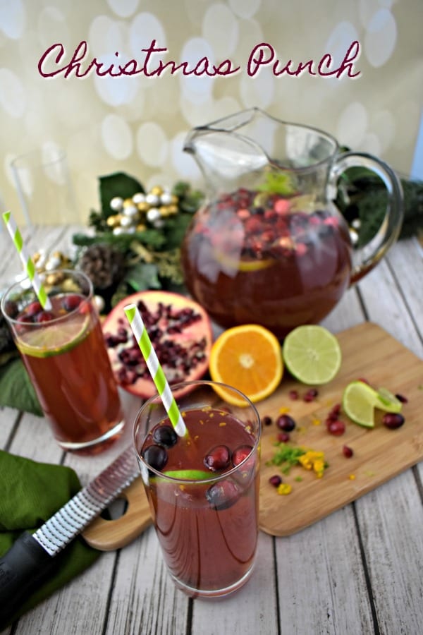 Pomegranate Punch in a glass pitcher along with tall glasses of punch garnished with fresh limes, oranges, cranberries and pomegranate seeds.