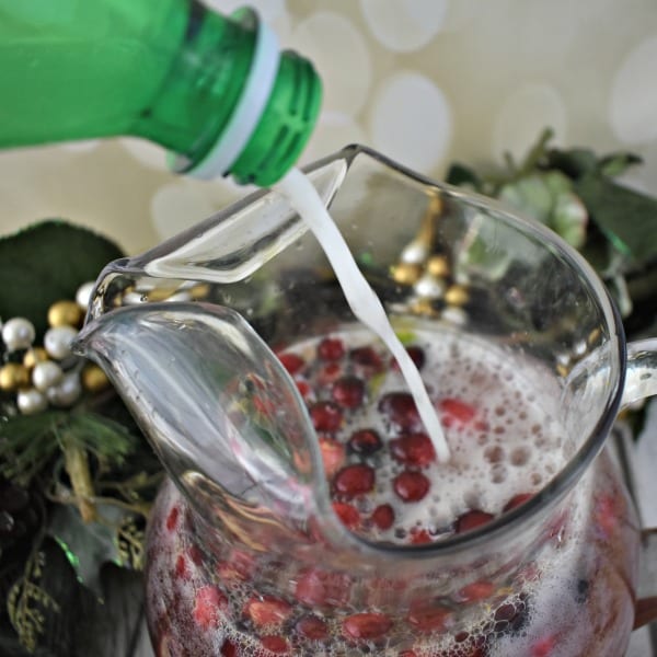 Soda pours into a glass pitcher with other liquids and fresh cranberries.