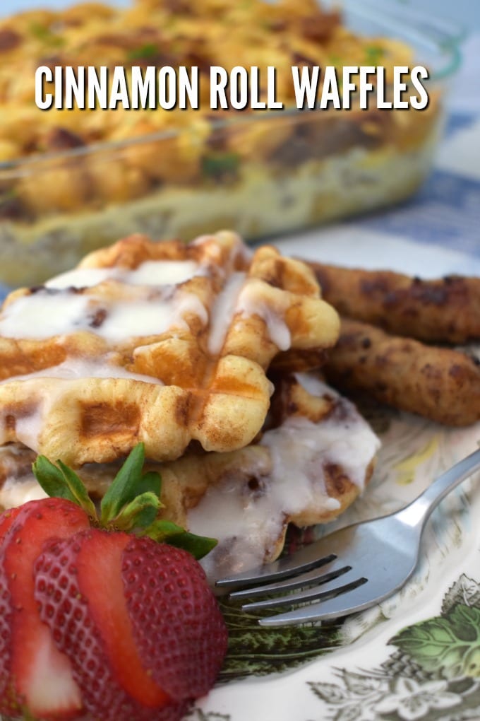 Cinnamon Rolls Waffles drizzled with icing with breakfast sausage and a cut strawberry and a fork. A breakfast casserole sits in the background.