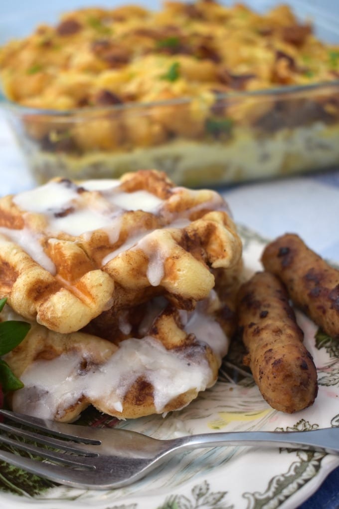 Cinnamon Roll Waffles drizzled with icing with breakfast sausage and a fork. A breakfast casserole sits in the background.