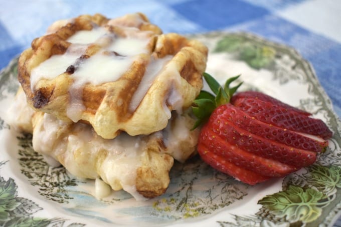 Two cinnamon waffles on a plate with a cut strawberry on a Friendly Village china plate set on top of a blue and white checked cloth.