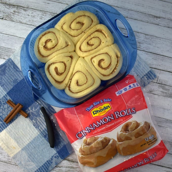 A baking dish with cinnamon roll dough sits on a table with a package of rolls and a blue and white checked towel.