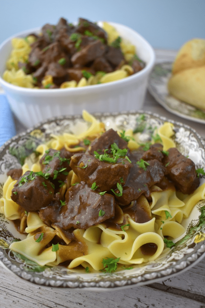 A bowl of beef tips and gravy over eggnoodles is shown with dinner rolls and a serving dish in the background.