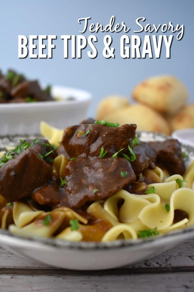 A bowl of beef tips and gravy over eggnoodles is shown with dinner rolls and a serving dish in the background.