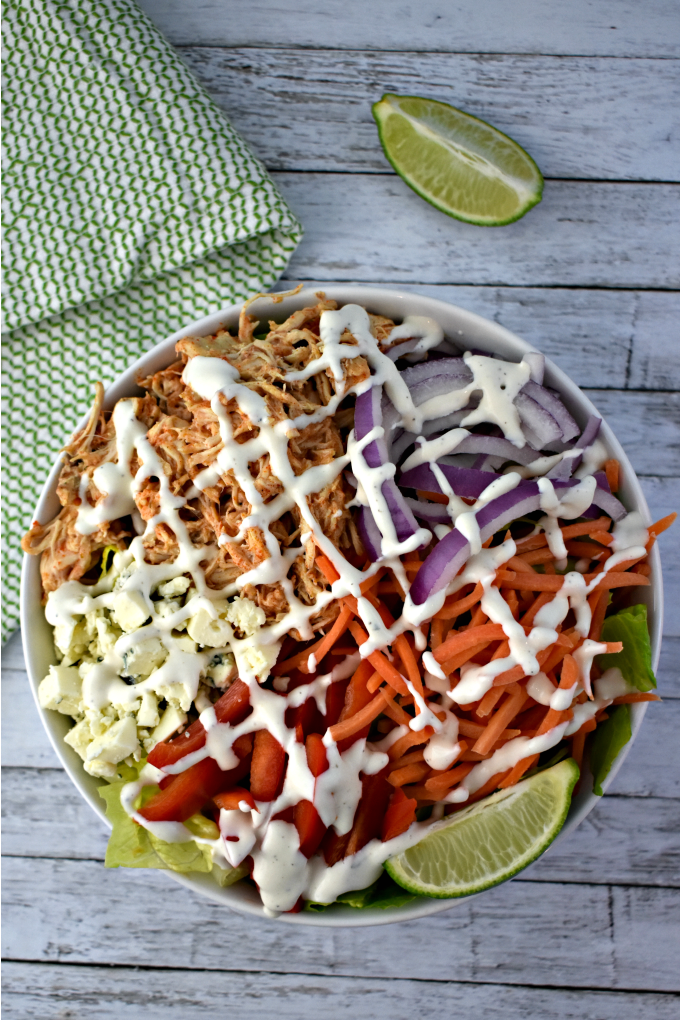 A large white bowl of buffalo chicken salad including crumbled blue cheese, shredded carrots, thinly sliced red onions, chopped tomatoes drizzled with ranch dressing sits on a white table near a wedge of lime and a green and white kitchen towel.