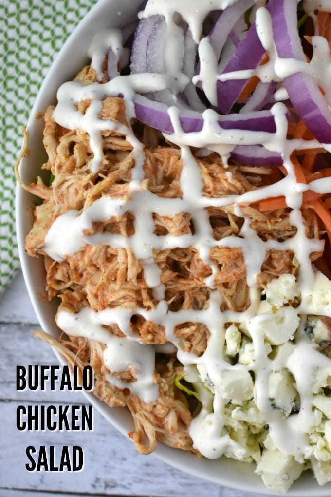 A close of a large white bowl of buffalo chicken salad including crumbled blue cheese, shredded carrots, thinly sliced red onions, chopped tomatoes drizzled with ranch dressing sits on a white table near a green and white kitchen towel.