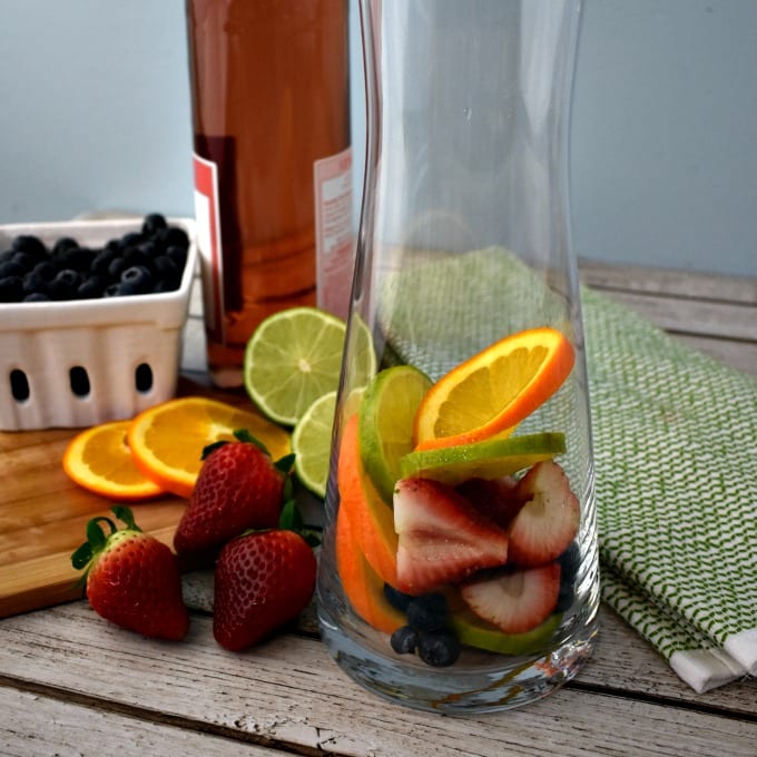 Glass pitcher filled with fruit on wooden cutting board.