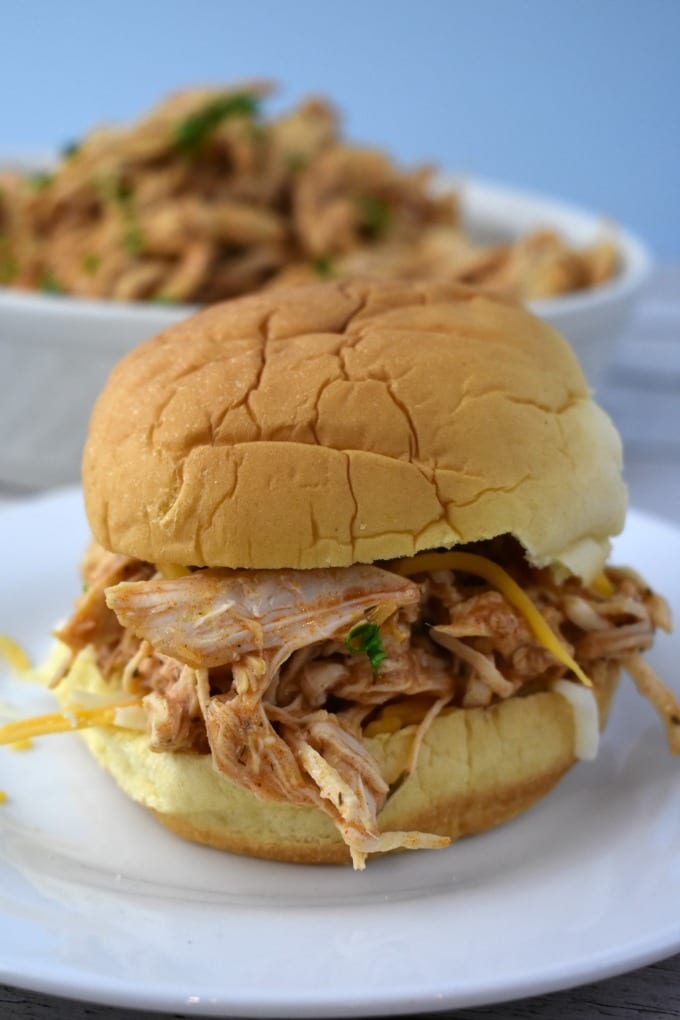 A slow cooker buffalo chicken sandwich on a white plate with a bowl of shredded buffalo chicken in a bowl in the background.