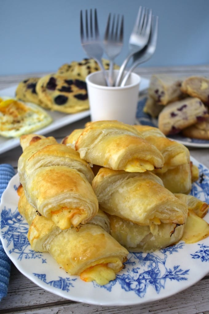 Ham and cheese croissants piled on a blue and white plate in front of a brunch spread.