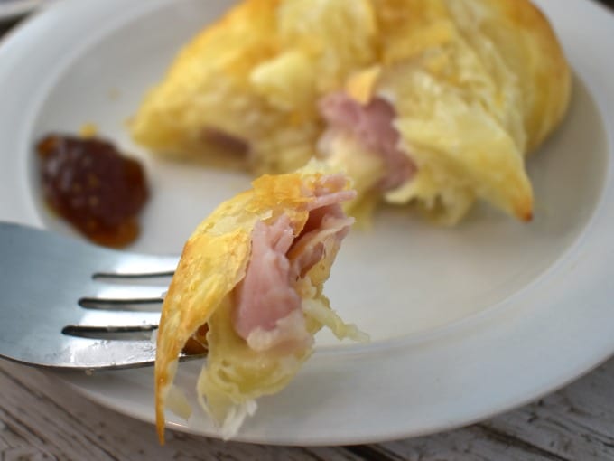 A ham and cheese croissant on a white plate with fig jam. One bite of the croissant is on a silver fork.