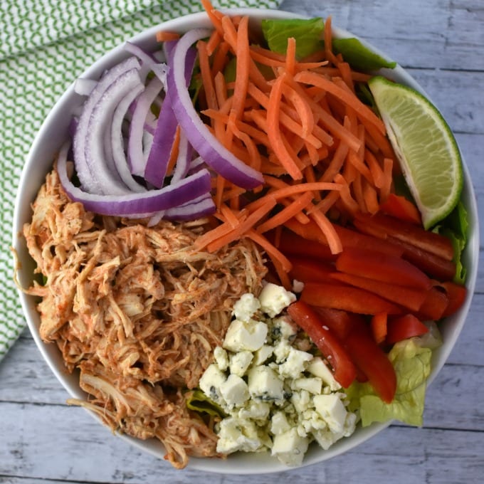 A large white bowl of Buffalo Chicken Salad including slow cooked buffalo chicken, shredded carrots, thinly sliced red onion, sliced red peppers, a wedge of lime and crumbled blue cheese sits on a white table with a green and white kitchen towel.