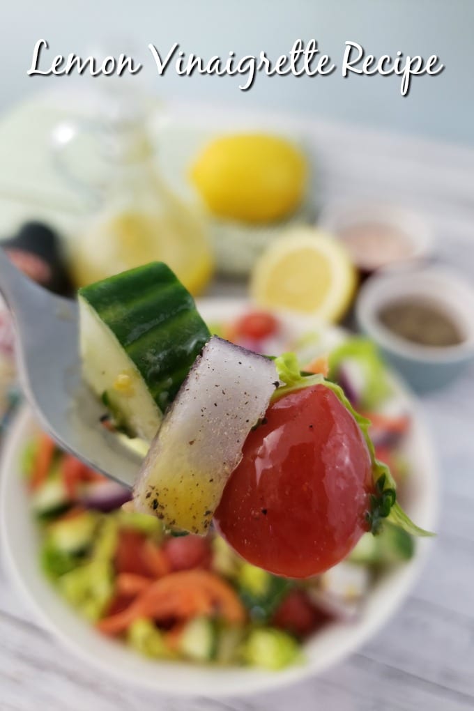 A forkful of garden salad with Lemon Vinaigrette dripoing from it in the foreground with a garden salad, dressing cruet and fresh lemon in the background