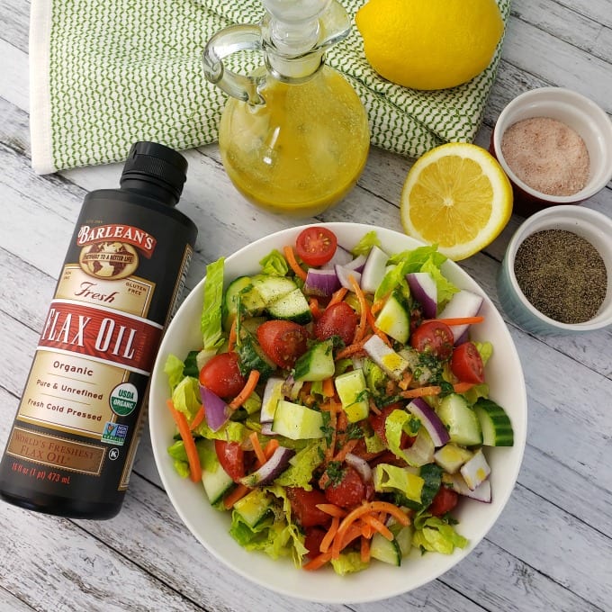 A bowl of garden salad sits on a table with a bottle of Barleans Flax Oil, a cruet of lemon dressing, a whole lemon and sliced lemon, and ramekins of salt and pepper.