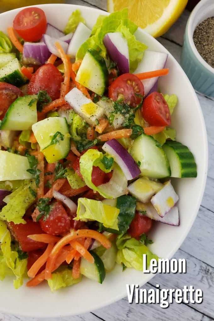 A garden ssalad with cucumbers, tomatoes and carrots drizzled with Lemon Vinaigrette. A halved lemon and ramekins with salt and pepper.