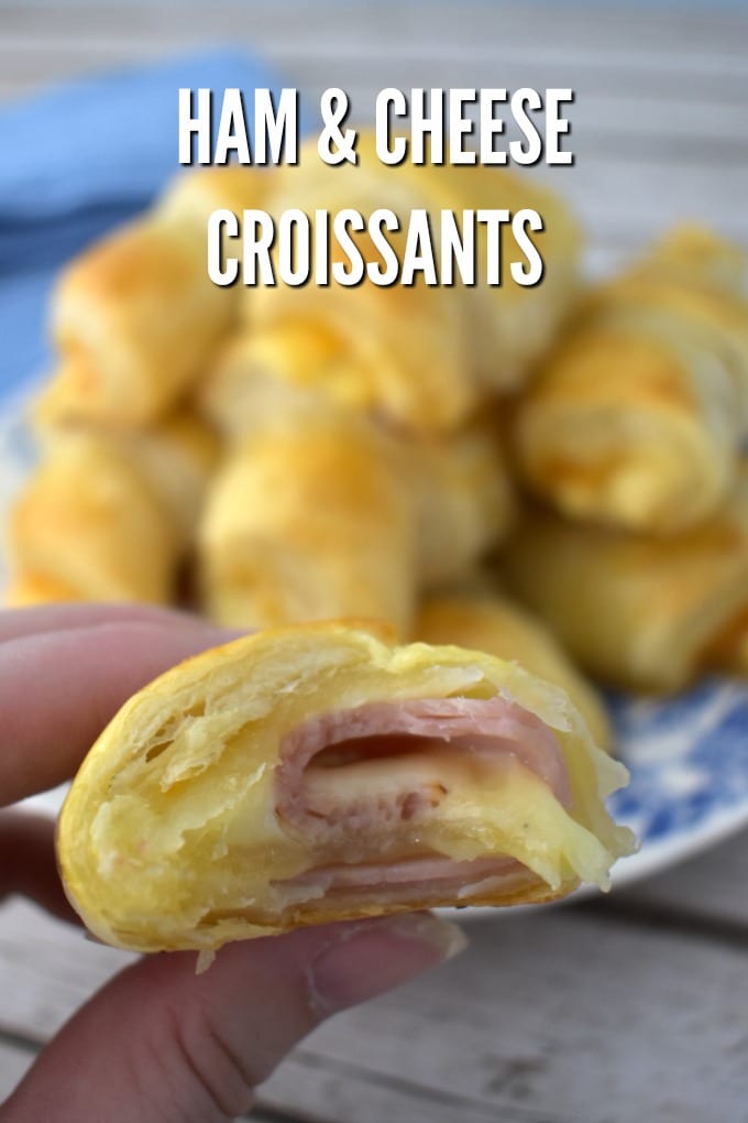 A hand holidng a ham and cheese croissant with a bite taken out of it. A blue and white plate full of whole croissants sits in the background.