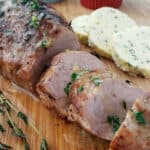 Slices of oven roasted pork tenderloin on a cutting board with a sprig of fresh thyme. In the background are slices of compound butter and ramekins with salt and pepper.