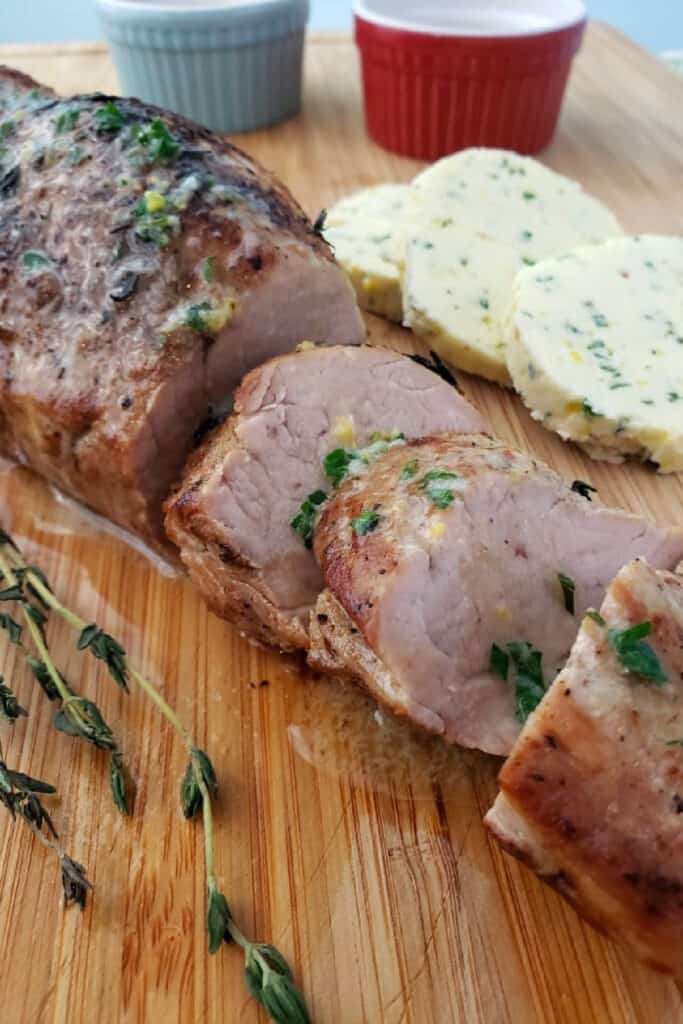 Slices of oven roasted pork tenderloin on a cutting board with a sprig of fresh thyme. In the background are slices of compound butter and ramekins with salt and pepper,