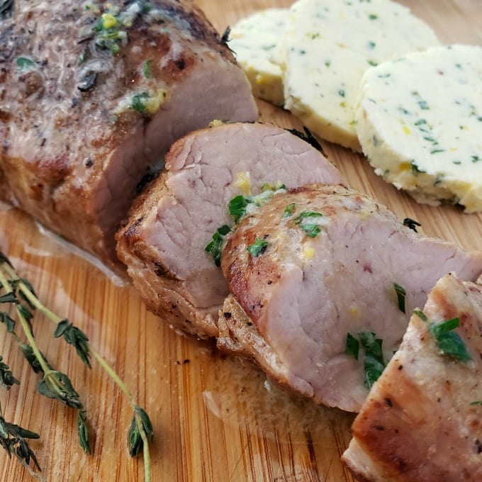 Slices of oven roasted pork tenderloin on a cutting board with a sprig of fresh thyme. In the background are slices of compound butter and ramekins with salt and pepper