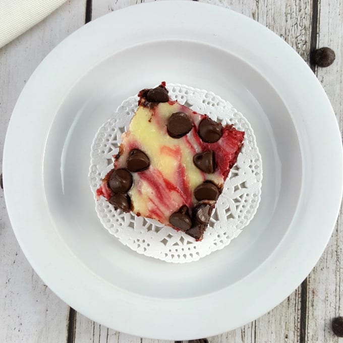 Arial view of a red velvet cheesecake brownie with chocolate chips on a white doily placed on a white plate. The plate is on a white wood background and there is a single chocolate chip on the table next to the plate.