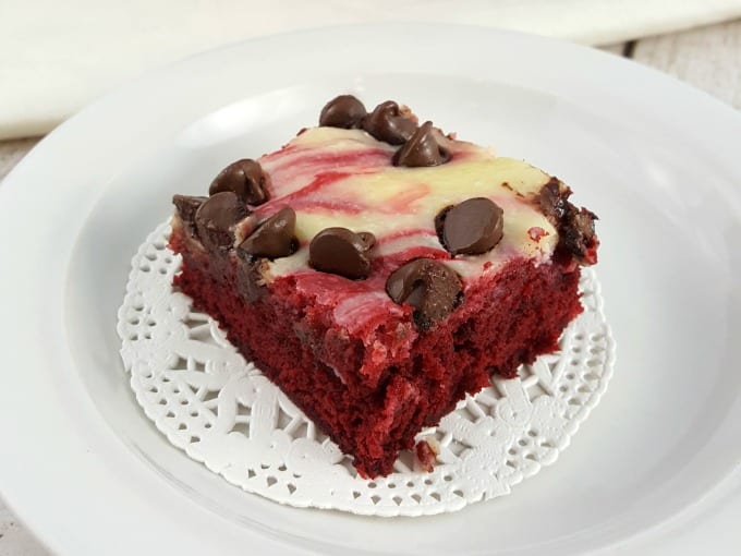 Close up of a red velvet cheesecake brownie with chocolate chips sitting on a white doily and on a white plate.